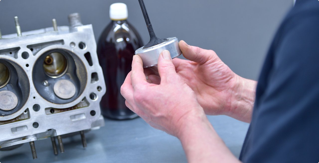 A close-up of a person's hands holding a tool, possibly a measuring device, with an engine cylinder head featuring exposed valves and pistons visible in the background, alongside a bottle of dark liquid, suggesting mechanical work or engine maintenance.