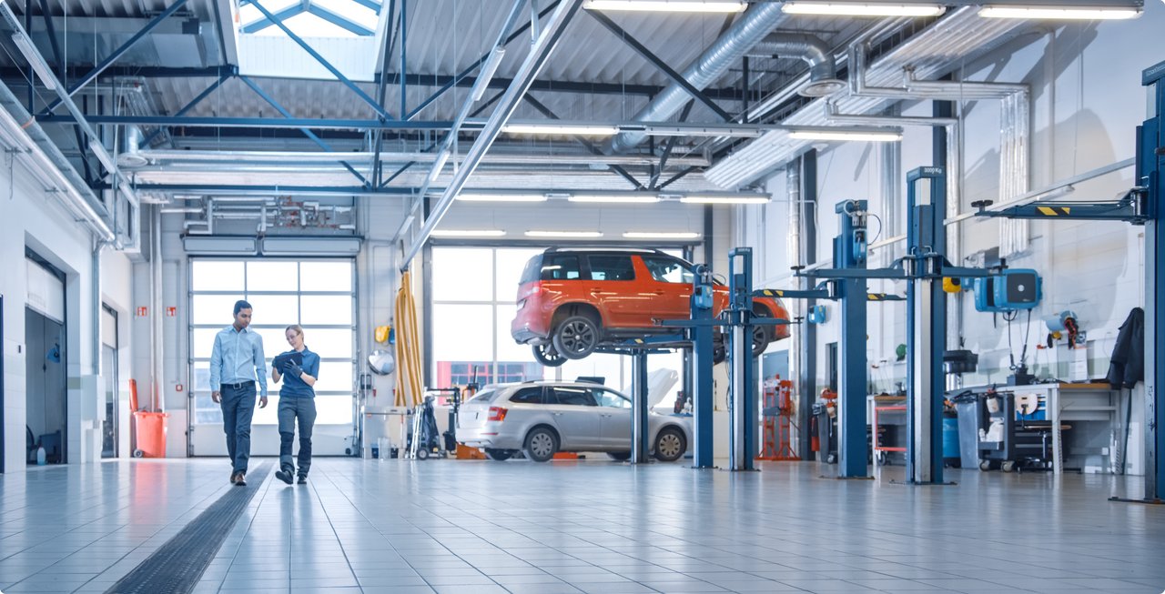 A modern car workshop with two mechanics walking and talking in the foreground. In the background, several car lifts are visible, one with an orange vehicle on it and a silver car beneath it.