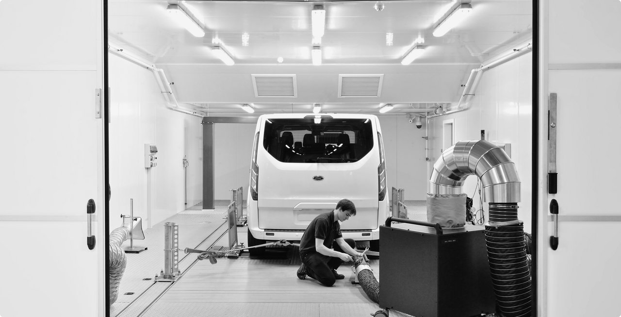 Man working in the rear of a white van parked inside a hall, surrounded by measurement equipment.