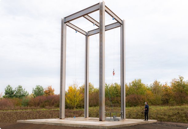 A tall, open steel structure in the form of a cube or frame stands outdoors on a pedestal, with a person visible beside it. In the background are trees and a cloudy sky.