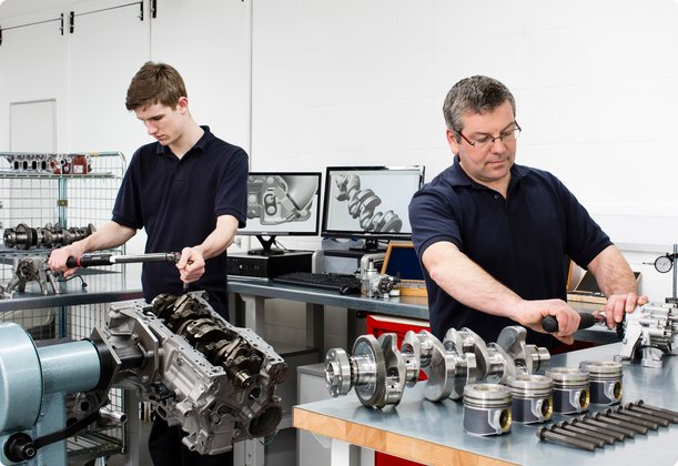Two technicians or mechanics, one younger and one older, are focused on working with engine components and other precise parts in a clean workshop or laboratory.