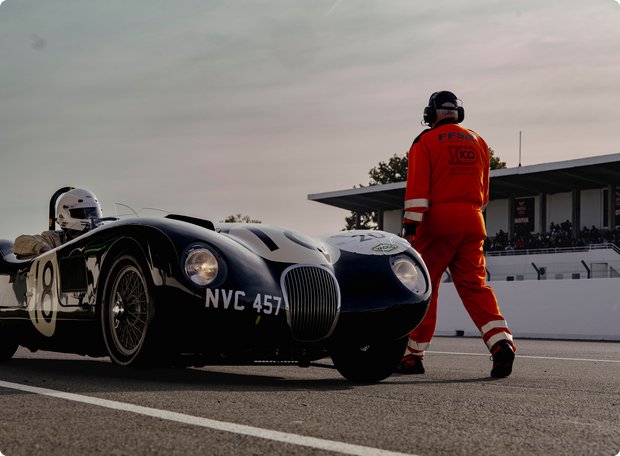 A mechanic or race team member in a red racing suit and helmet stands next to a classic black race car on a race track. A second race car is faintly visible in the background.