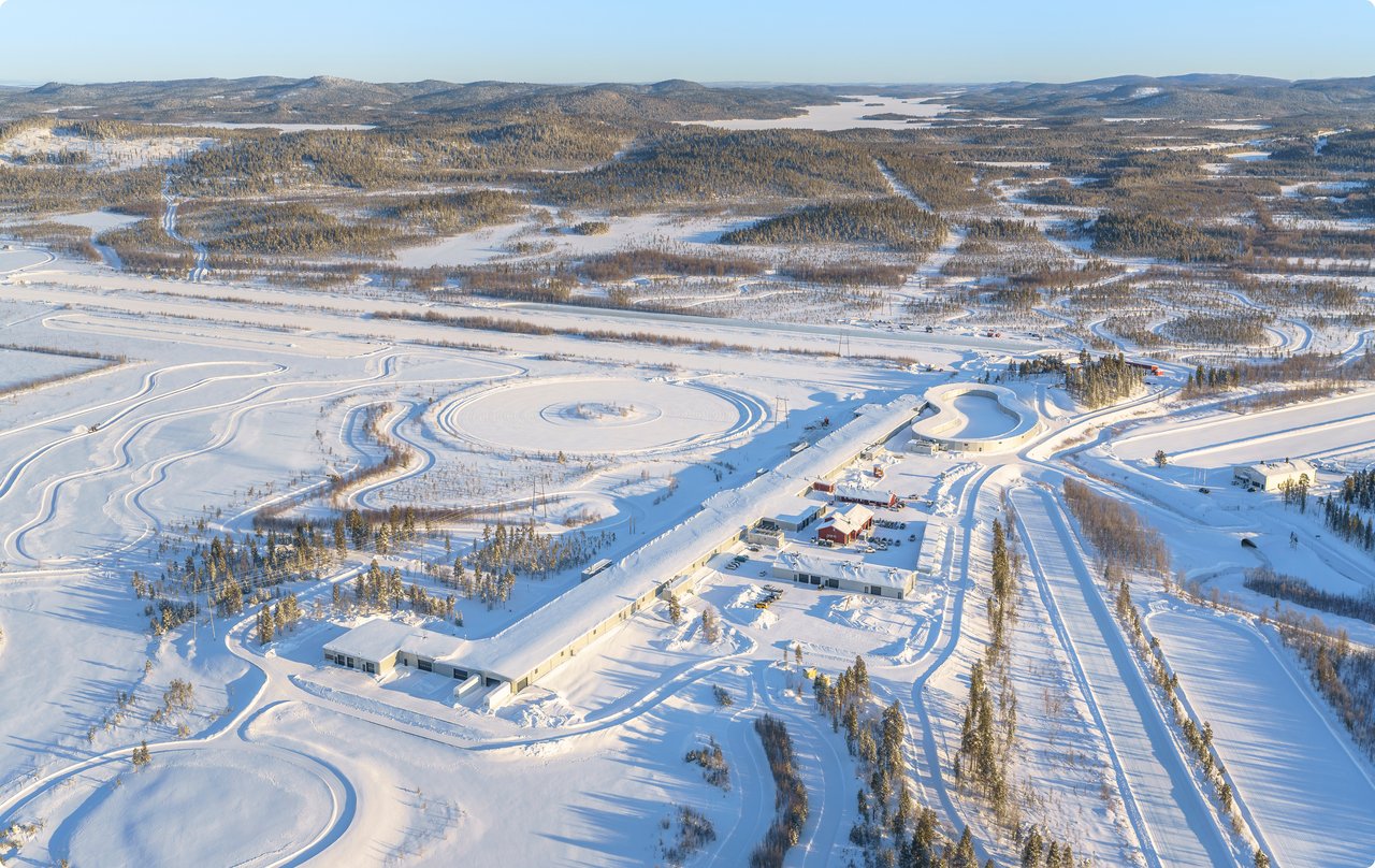 A vast winter landscape, captured from a bird's-eye view, featuring a large, snow-covered open area in the foreground bordered by rows of trees and a snow-covered settlement or town in the background. Some tracks or structures are discernible in the snow.