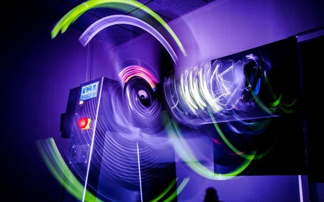 An abstract, dynamic photograph of a light sculpture or installation, showing rapidly rotating or vibrating lights in purple, pink, and neon green, leaving light trails in the air. The scene is dark, emphasizing the glowing colors and the movement of the lights.