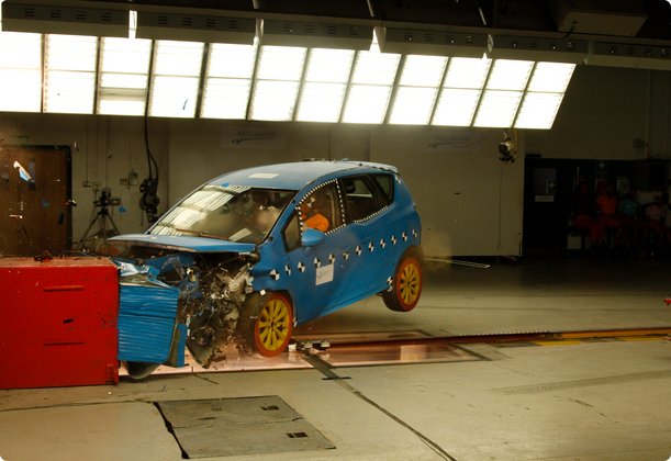A blue compact car in the midst of a frontal crash test, impacting a red barrier. The front of the vehicle is severely deformed, and the car is slightly lifted off the ground, capturing the moment of impact.
