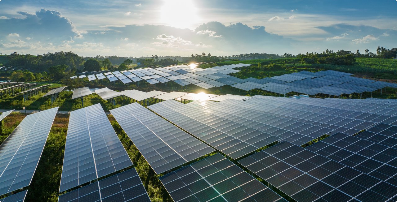 A sprawling aerial shot of a solar farm with countless photovoltaic panels bathed in bright sunlight. The sun reflects off some of the panels, and in the background, green vegetation, trees, and a cloudy sky are visible. The scene symbolizes renewable energy.