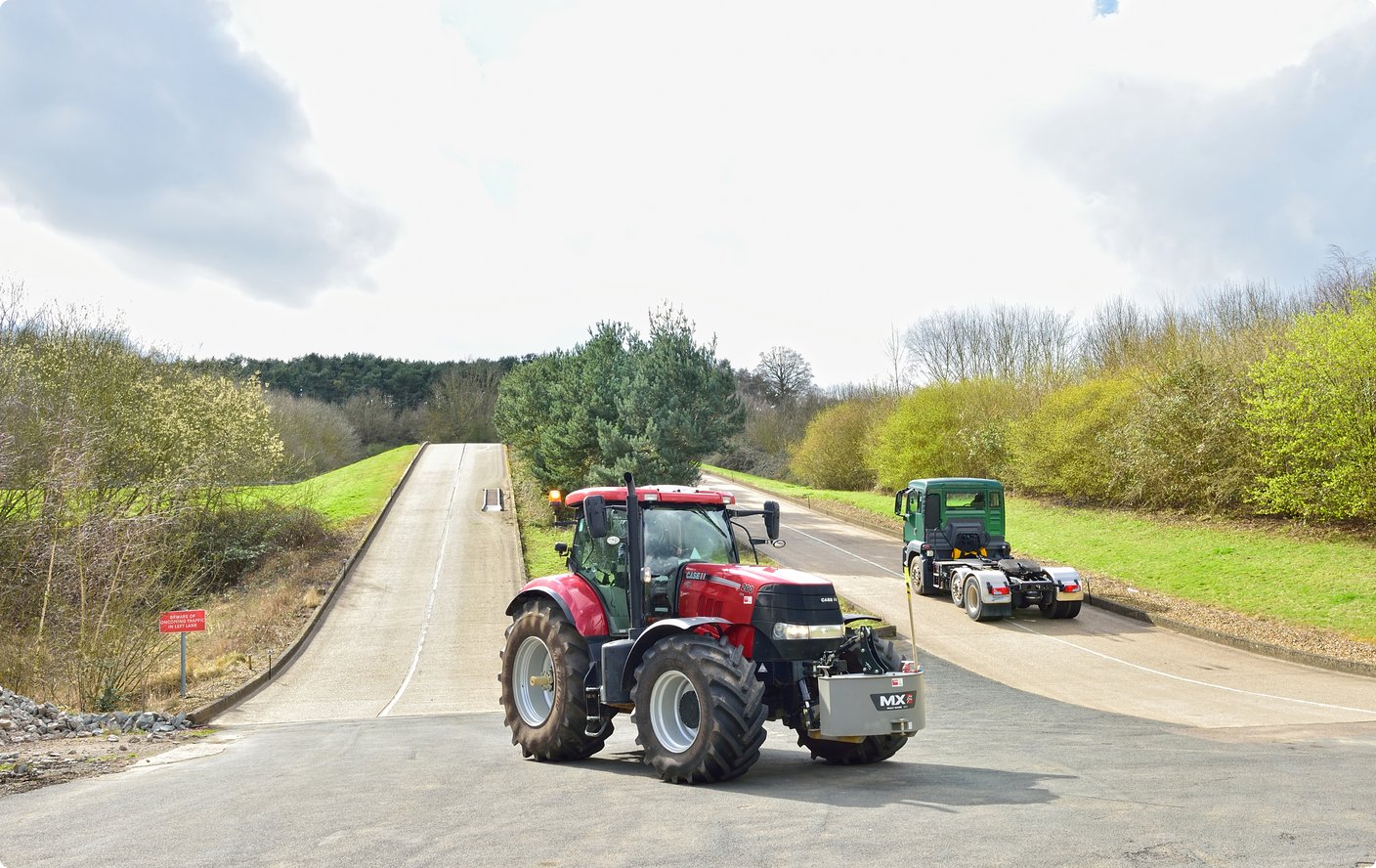 Ein roter Traktor steht auf einem Schotterweg in einer landwirtschaftlichen Umgebung, mit einem weiteren kleineren grünen landwirtschaftlichen Gerät oder Anhänger dahinter. Üppige grüne Felder und Bäume säumen den Weg unter einem teilweise bewölkten Himmel.