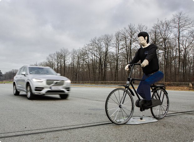 A test scene on a road, showing a silver SUV approaching a stationary test dummy on a bicycle. The scene takes place under a cloudy sky with trees in the background.