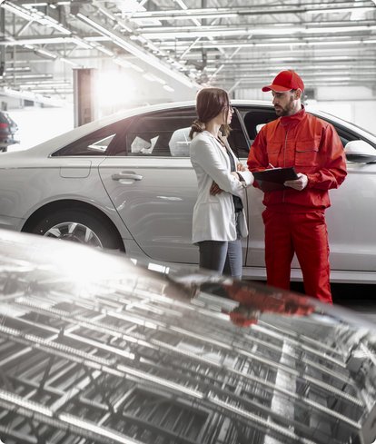 A mechanic in a red overall and cap consults with a female customer or colleague in a car dealership or workshop, with a white car in the background.