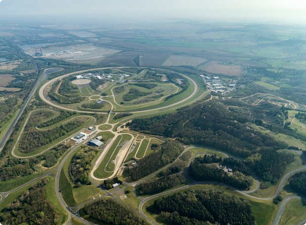 A sprawling aerial view shows a vast test track or proving ground, composed of multiple interconnected roads and oval courses, surrounded by forests and open fields under a wide sky.
