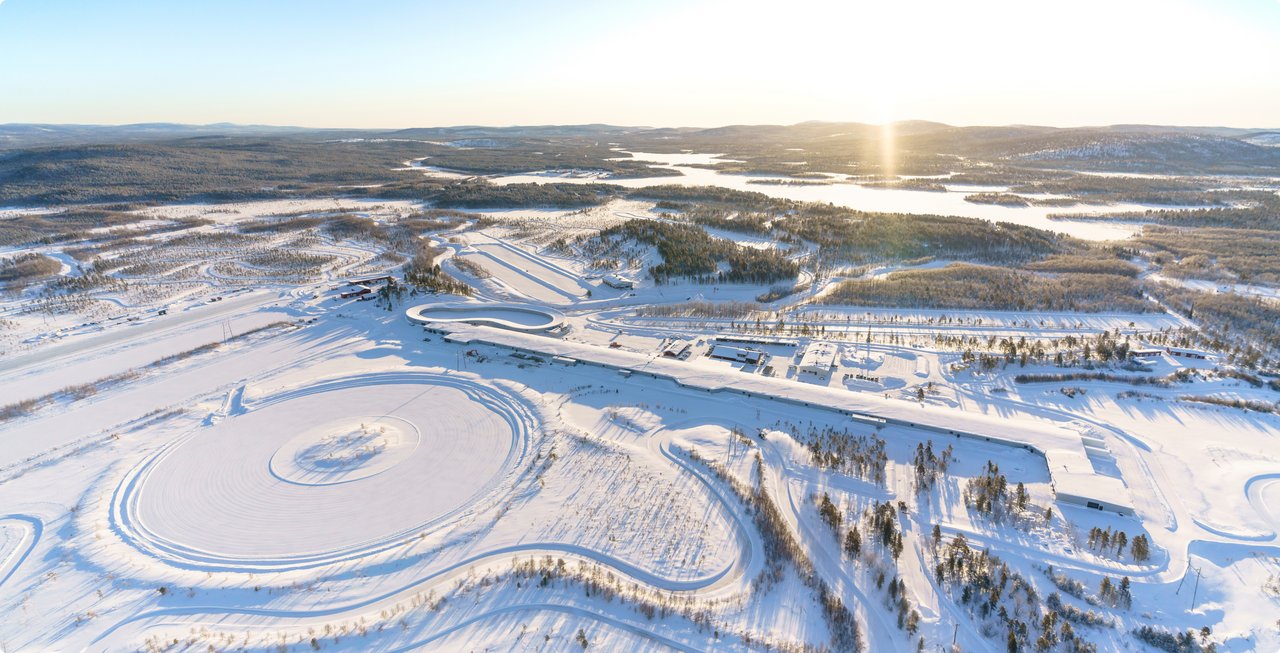 An aerial view of a vast, snow-covered landscape with a frozen lake or body of water reflecting the bright sunlight in the distance. The foreground shows a complex network of snow tracks and cleared areas, likely for winter testing or activities.
