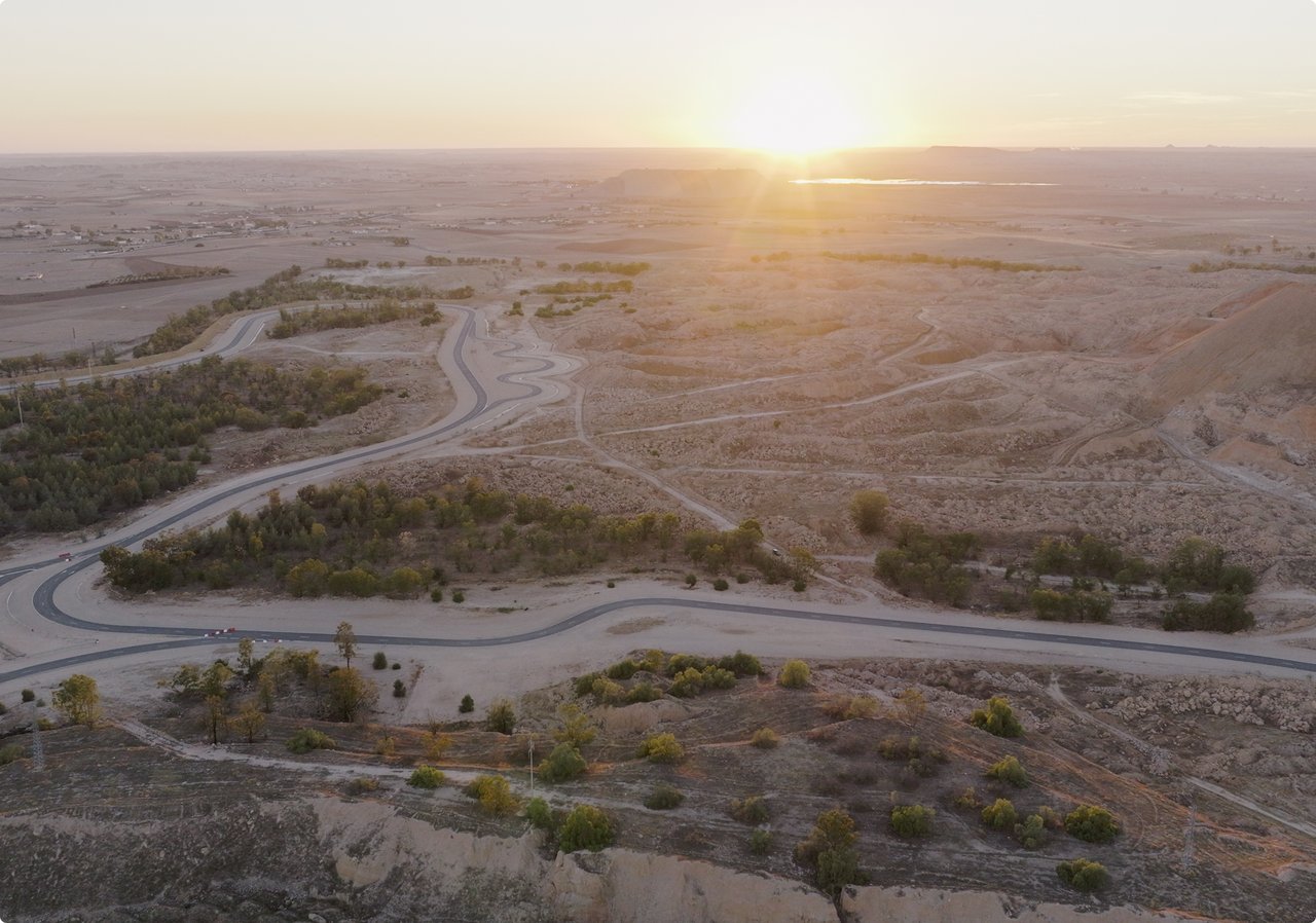 A vast, arid desert landscape at sunset, featuring narrow green oases and winding paths. The sun is low on the horizon, casting a warm glow.
