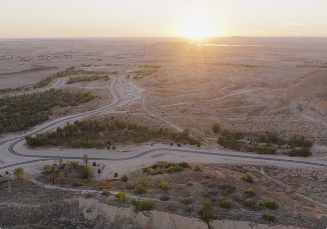 Un vaste paysage désertique aride au coucher du soleil, avec d'étroites bandes d'oasis vertes et des chemins serpentant à travers le terrain. Le soleil est bas à l'horizon, projetant une lueur chaleureuse.