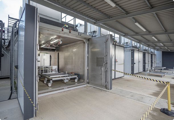An open, large, walk-in metal chamber with technical equipment inside, part of a series of similar closed chambers in an industrial hall.