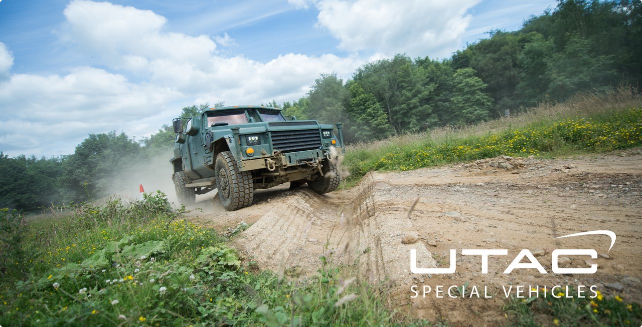 An off-road military vehicle drives on an unpaved dirt track through a rural or wooded area. The vehicle is in dark colors and kicks up dust. The UTAC logo is visible in the lower right area of the image.