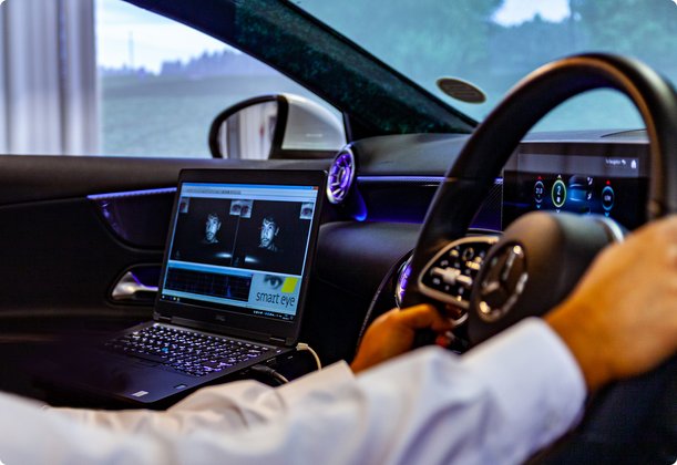 A driver in a shirt, with hands on the steering wheel, sits inside a car, with a laptop on the dashboard likely used for testing or data recording.