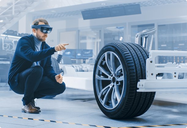 An engineer or technician wearing augmented reality glasses squats in a modern factory hall or laboratory, interacting with a virtual 3D model of a car tire and chassis.