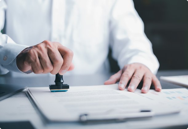 A close-up shot shows a person in a white shirt stamping an official document or form with a rubber stamp, signifying approval, certification, or authentication.
