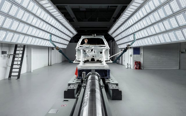 An empty car body on a test rig in a brightly lit laboratory or test track with rows of overhead lights.