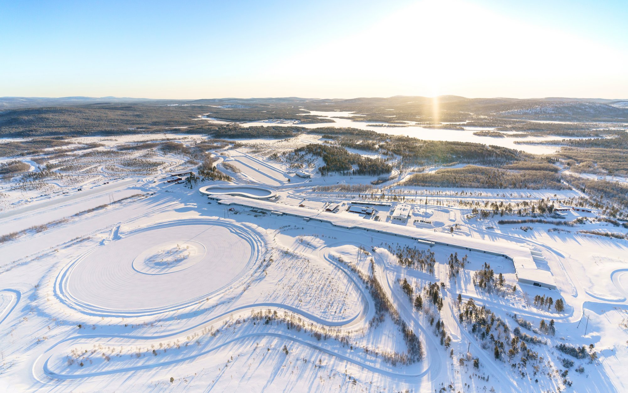 Eine weite Luftaufnahme eines verschneiten Testgeländes mit verschiedenen Fahrspuren und einem runden Testbereich, umgeben von Wäldern und zugefrorenen Seen. Die Sonne steht tief am Horizont.