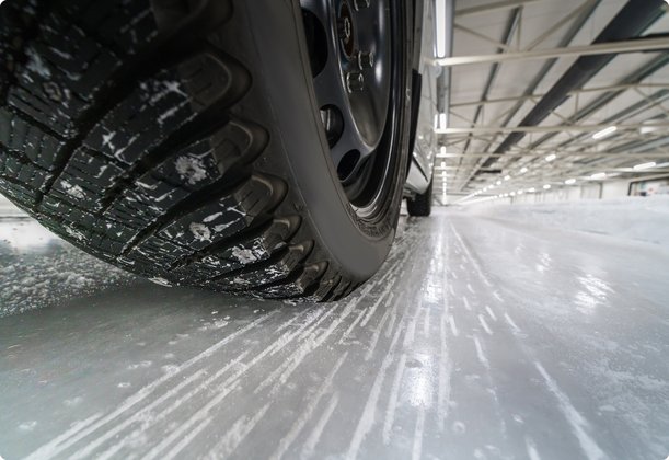 Close-up of a winter tire driving on a smooth, icy surface. Small ice fragments and grit particles are visible, being thrown off by the tire. A large, illuminated hall is recognizable in the background.