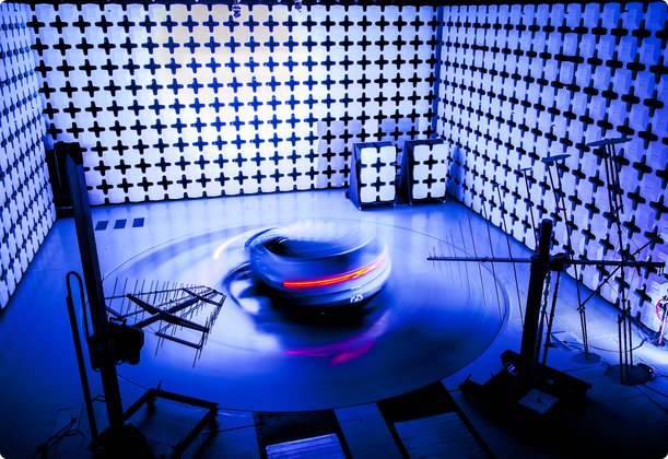An electromagnetically shielded chamber, also known as a Faraday cage or anechoic chamber, with a dark, low-profile vehicle or object on a rotating platform in the center. The room's walls are lined with pyramid-shaped, blue-tinted absorbing material, and the scene is bathed in cool blue and purple lighting, suggesting a test environment for electromagnetic compatibility (EMC) or radio frequency (RF) measurements.