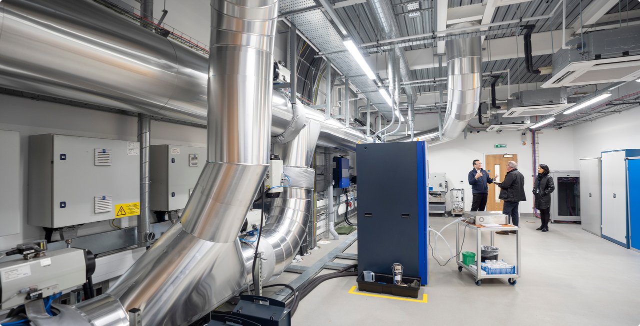A wide interior view of a modern industrial laboratory or facility featuring large, shiny metal pipes and complex machinery. Figures of people are faintly visible in the background, emphasizing the scale of the room.