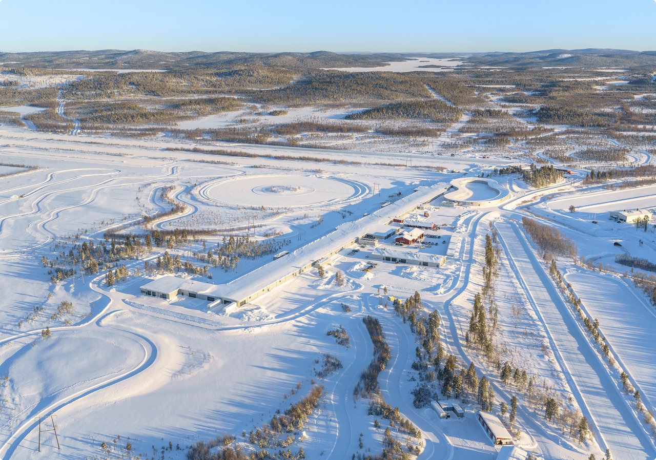 An aerial view of a snow-covered landscape with a long, white building or structure stretching through the center, flanked by what appear to be railway tracks and trees. In the background, a town or city is visible under a clear sky.
