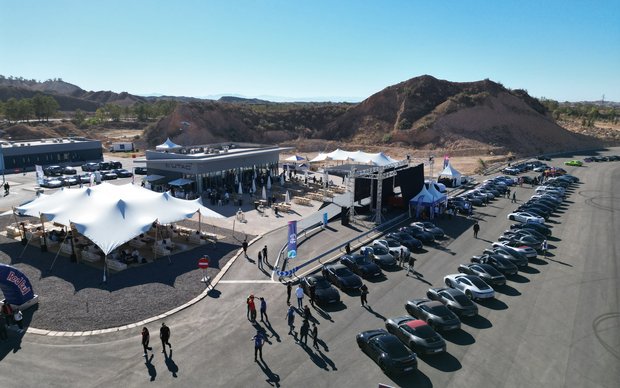 An aerial view of a large outdoor event or parking lot featuring several white tents, rows of black motorcycles, and some cars under a blue sky and mountainous landscape.