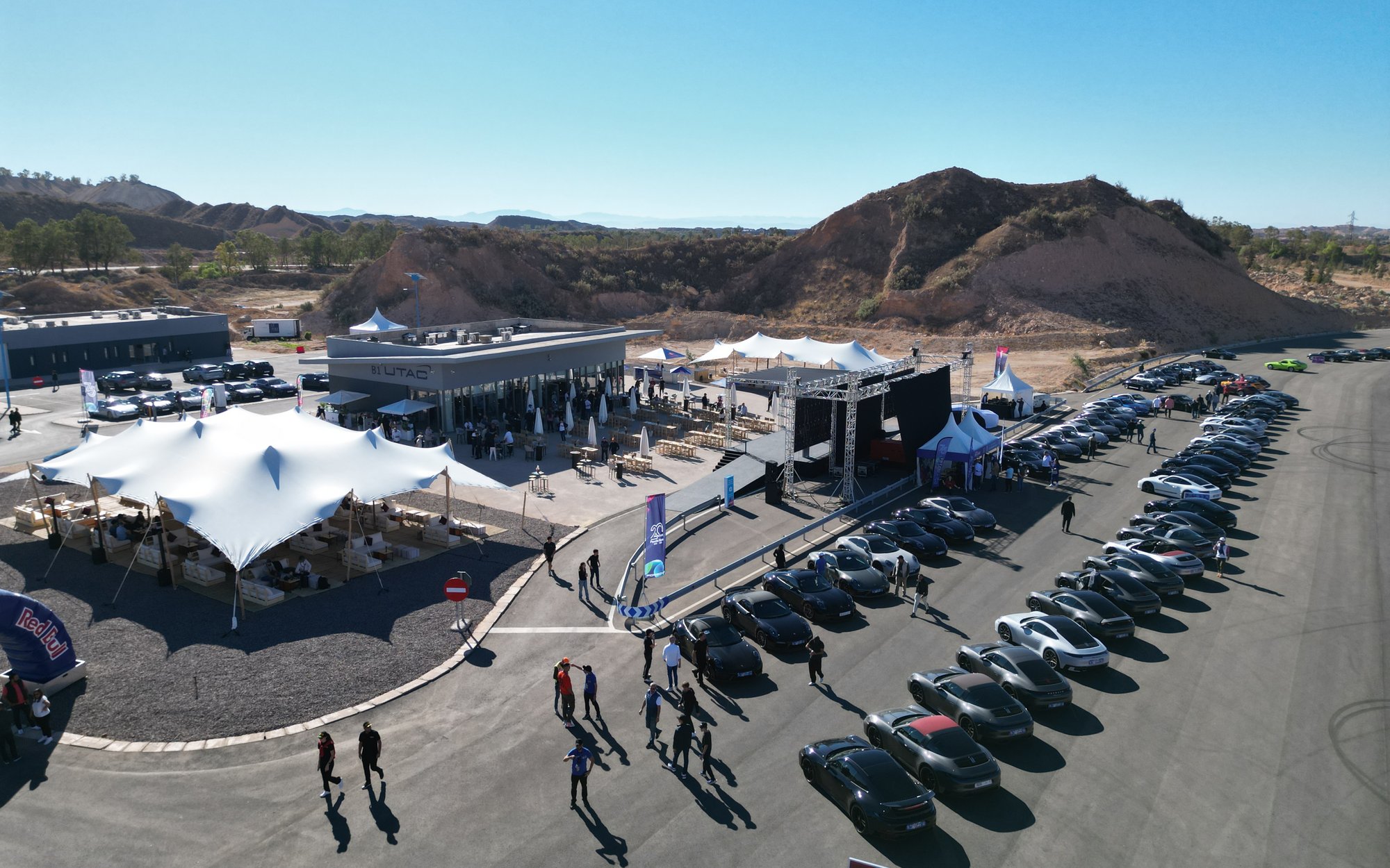 An aerial view of a large outdoor event or parking lot featuring several white tents, rows of black motorcycles, and some cars under a blue sky and mountainous landscape.