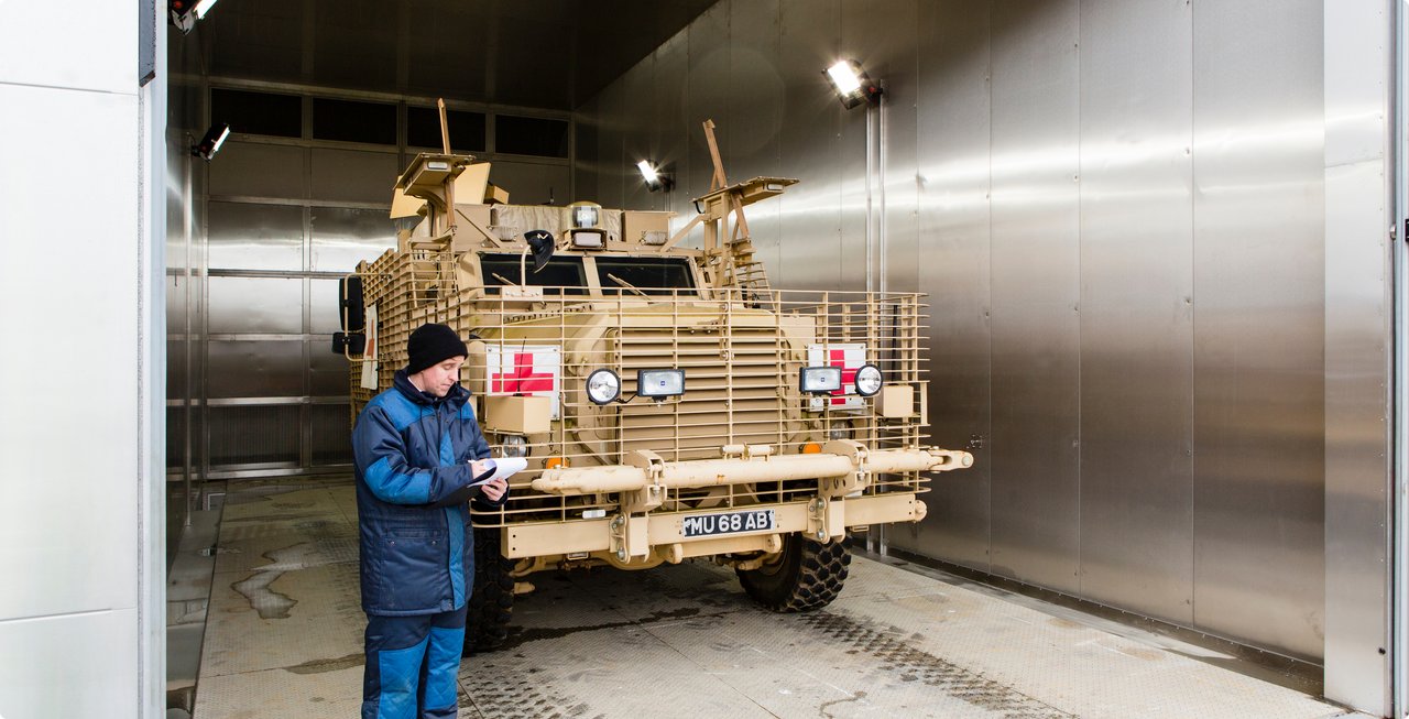 An armored military vehicle, possibly a prototype, is positioned in a large, cool or climate-controlled testing facility. A technician in warm clothing stands in front of it, taking notes.