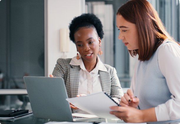 Two women are seated at a desk in a modern office, working collaboratively. One, a dark-skinned woman in a patterned jacket, sits in front of a laptop, while the other, a light-skinned woman, holds documents and looks at the screen, representing a collaborative discussion.