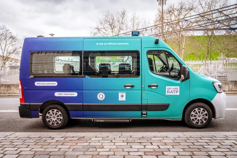 Side view of a modern van painted in a striking gradient from dark blue to turquoise. The vehicle is configured as a passenger transport or shuttle, with windows along the side and an elevated roof, possibly for sensors or additional equipment. It is parked on an urban street, with trees and buildings in the background.