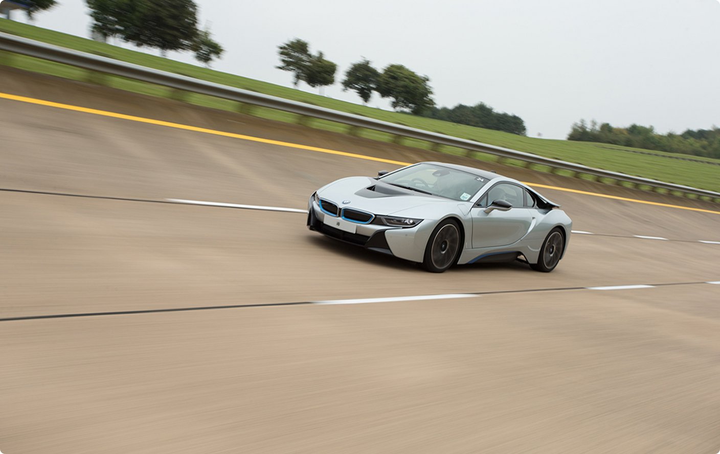 A sleek, silver sports car, possibly a BMW i8, is driving on a high-banked test track or circuit. The track surface is light-colored with dashed lines, and a guardrail with a yellow stripe runs along the top of the banking. Green trees and an overcast sky are visible in the background.