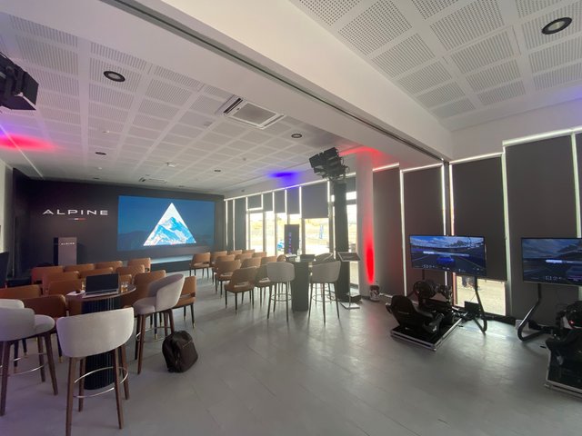 An empty conference or event room, prepared for an event. A large screen with a light blue triangle is visible in the background, surrounded by chairs and tables. The ceiling has a modern, perforated design, and lighting elements are visible.
