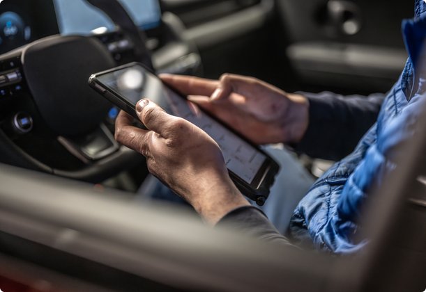 Close-up of a person's hands inside a car, holding and operating a smartphone in the left hand and a tablet in the right hand. The car's steering wheel is visible in the background.