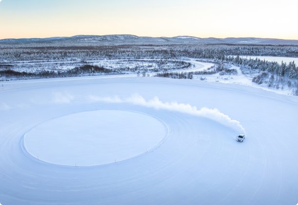 A drone shot of a vehicle driving in circles on a vast, snow-covered ice surface, leaving a trail of snow. In the background, snow-covered trees and a town are visible under a clear winter sky.