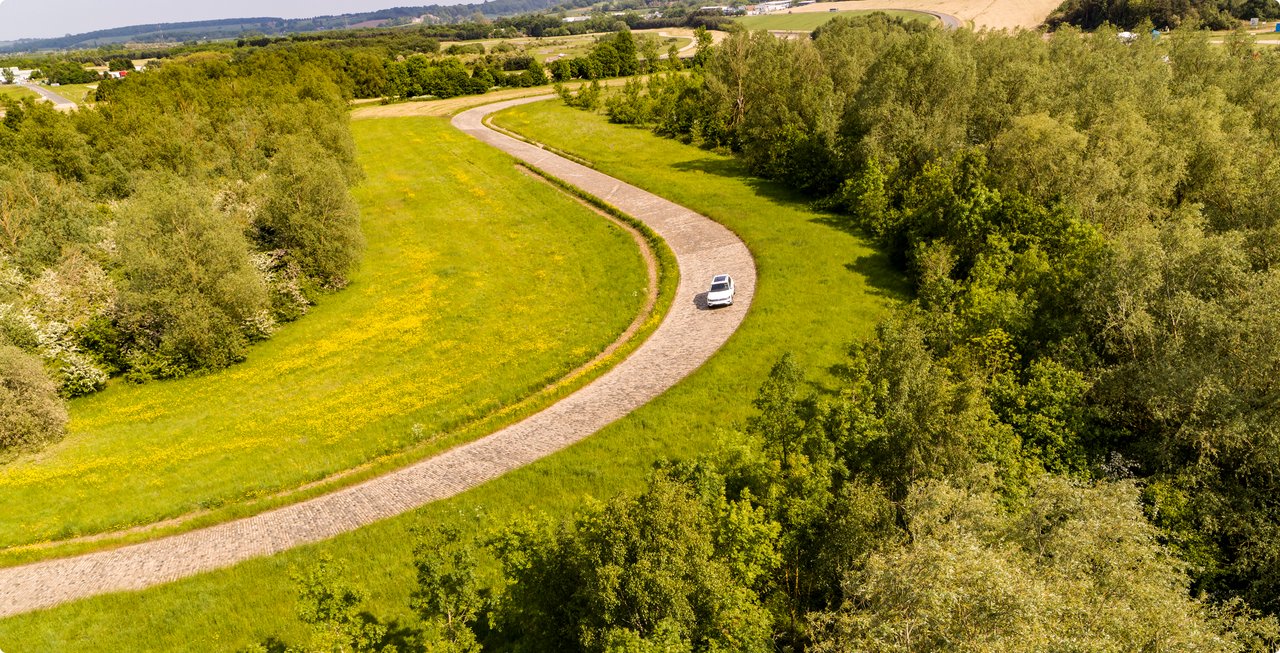 A white car drives on a winding road through a green forest and meadow landscape.