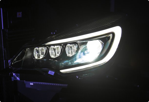 A close-up, dark shot of a modern car's headlight illuminated with bright white LED daytime running lights forming a distinctive shape around the main headlamp, which also emits a strong white light. The car's dark body is barely visible against the black background.