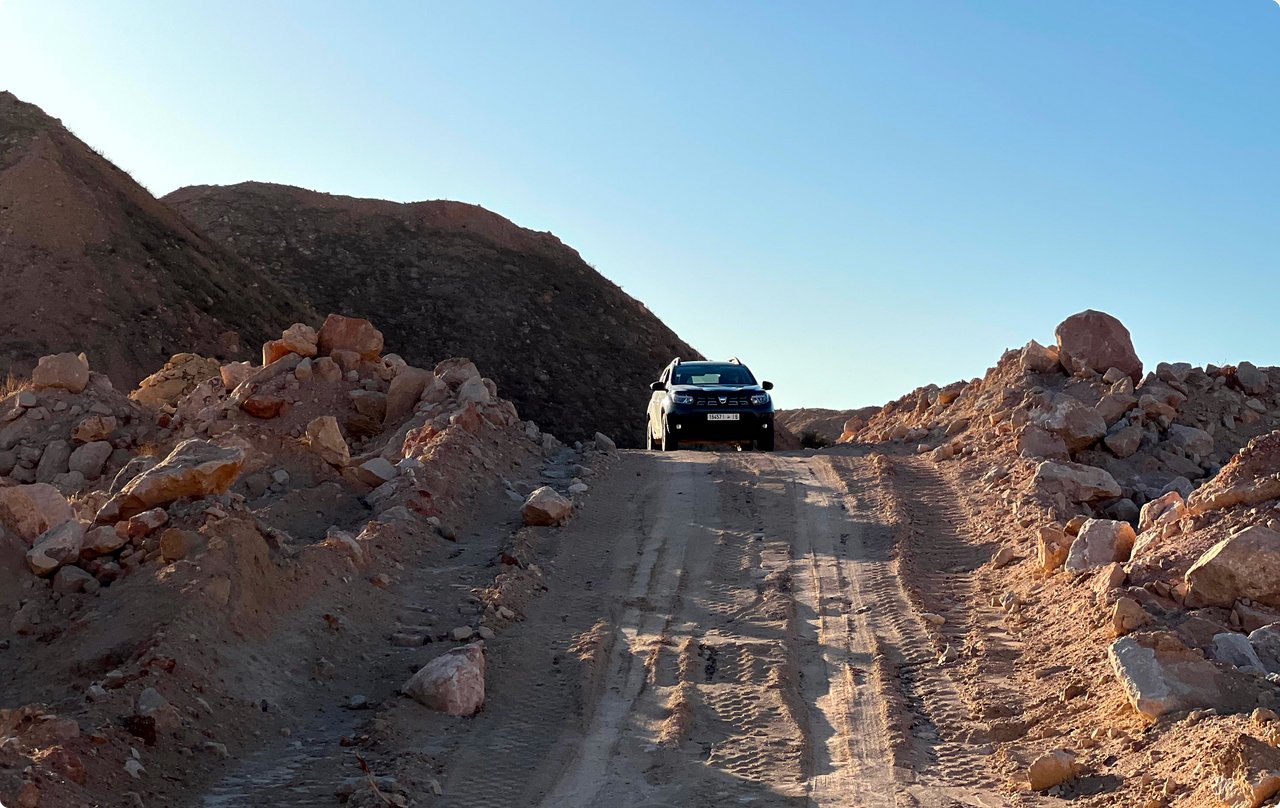 A dark vehicle, likely an SUV or off-road vehicle, is driving uphill on a steep, unpaved gravel path in a rocky, dry landscape. Large boulders line the path, and the sky is clear blue.