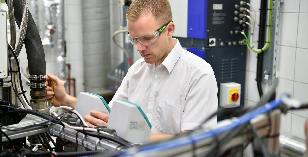A man wearing safety glasses and a white shirt is focused on working with complex machinery or equipment, holding documents or plans in his hands.