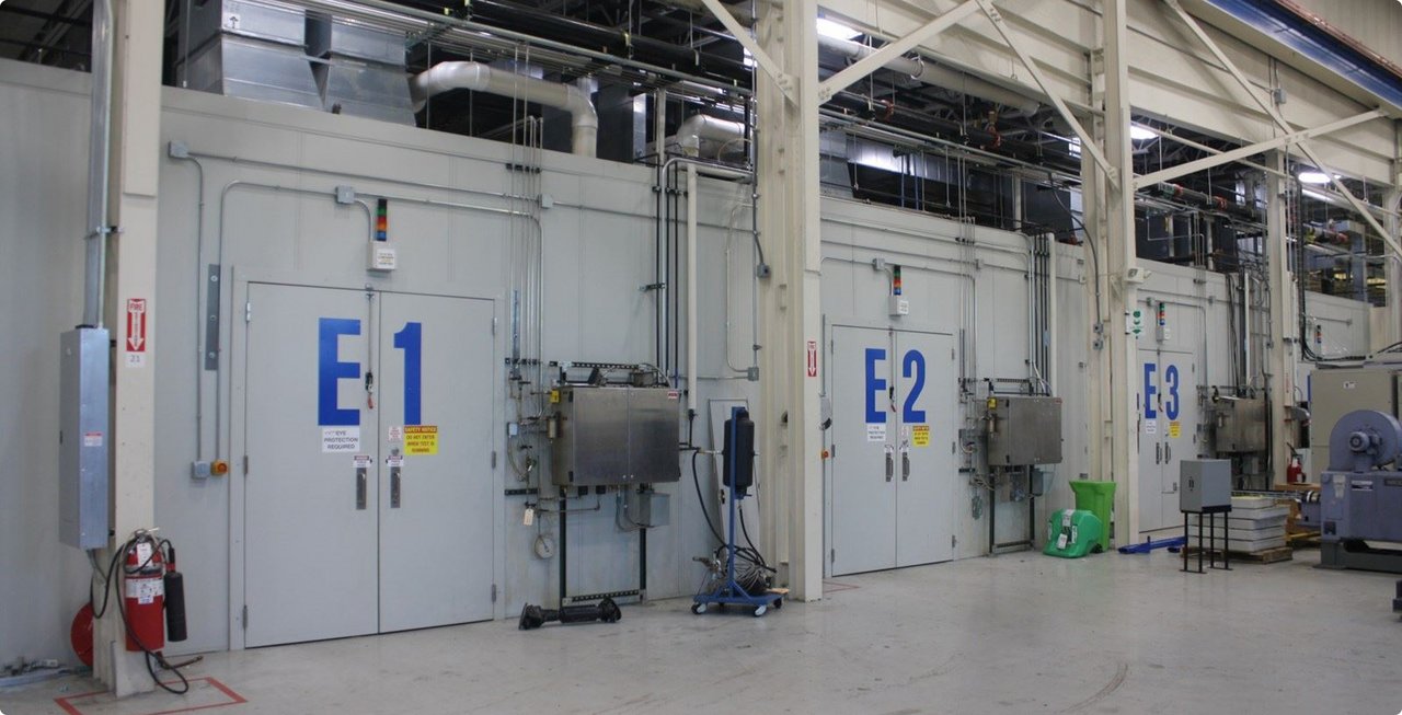 A series of large industrial test chambers or rigs in a factory hall. The chambers are grey and labeled with blue markings such as "E1", "E2", and "E3". Piping and technical equipment are visible.