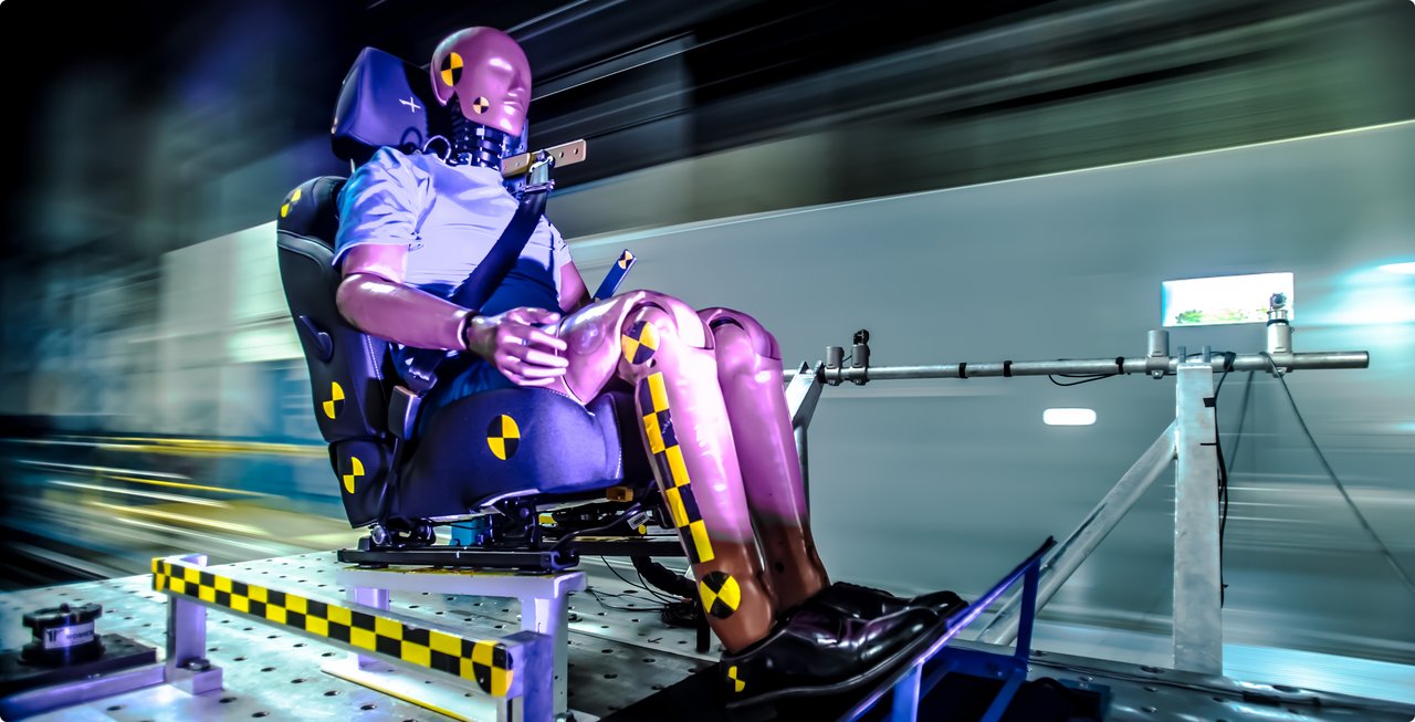 A crash test dummy with a seatbelt and sensors on its limbs, positioned on a seat in a test environment. The background is blurred, indicating motion or high speed.