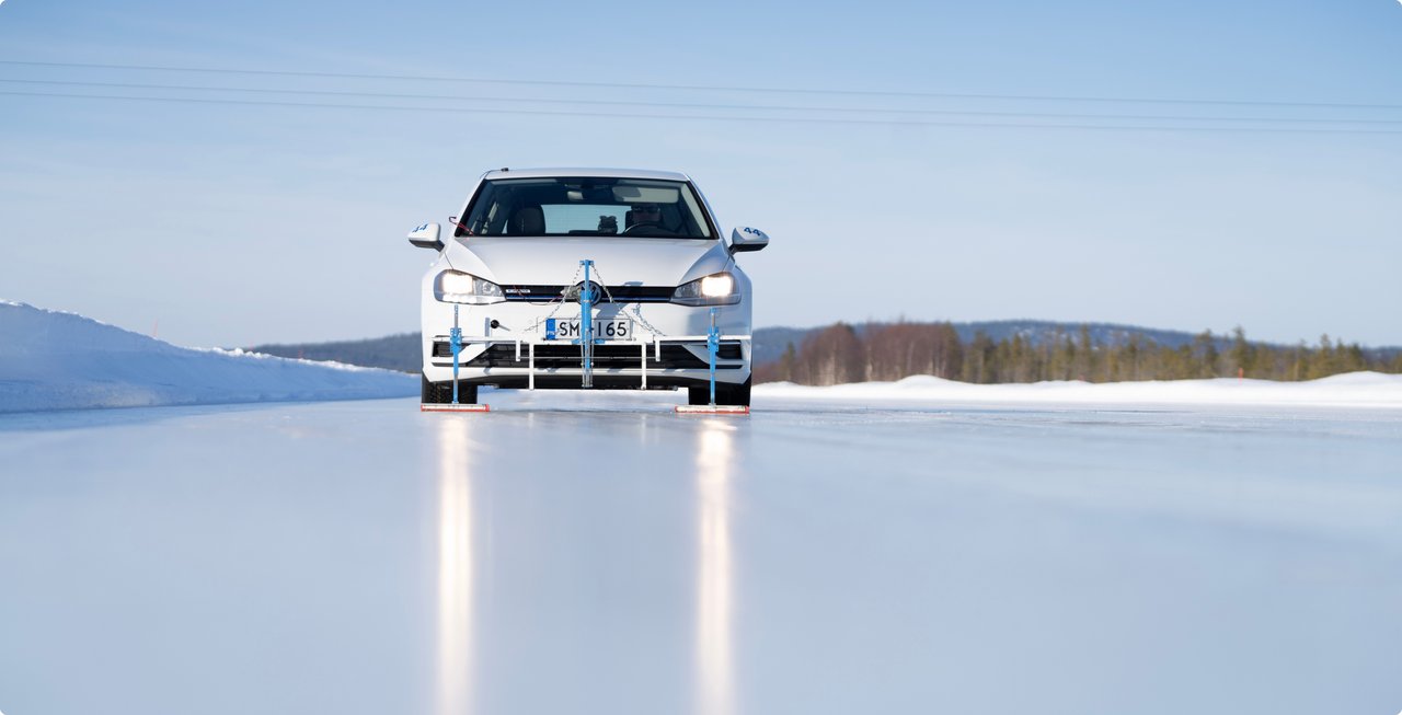 A white car drives on a vast, reflective icy surface, with its headlights casting reflections on the ice. Snow-covered trees are visible in the background.