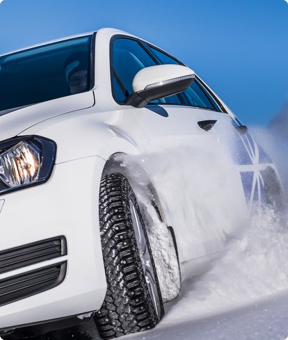 Close-up of the front of a white car with winter tires driving on a snowy or icy road, kicking up snow. The sky is blue.