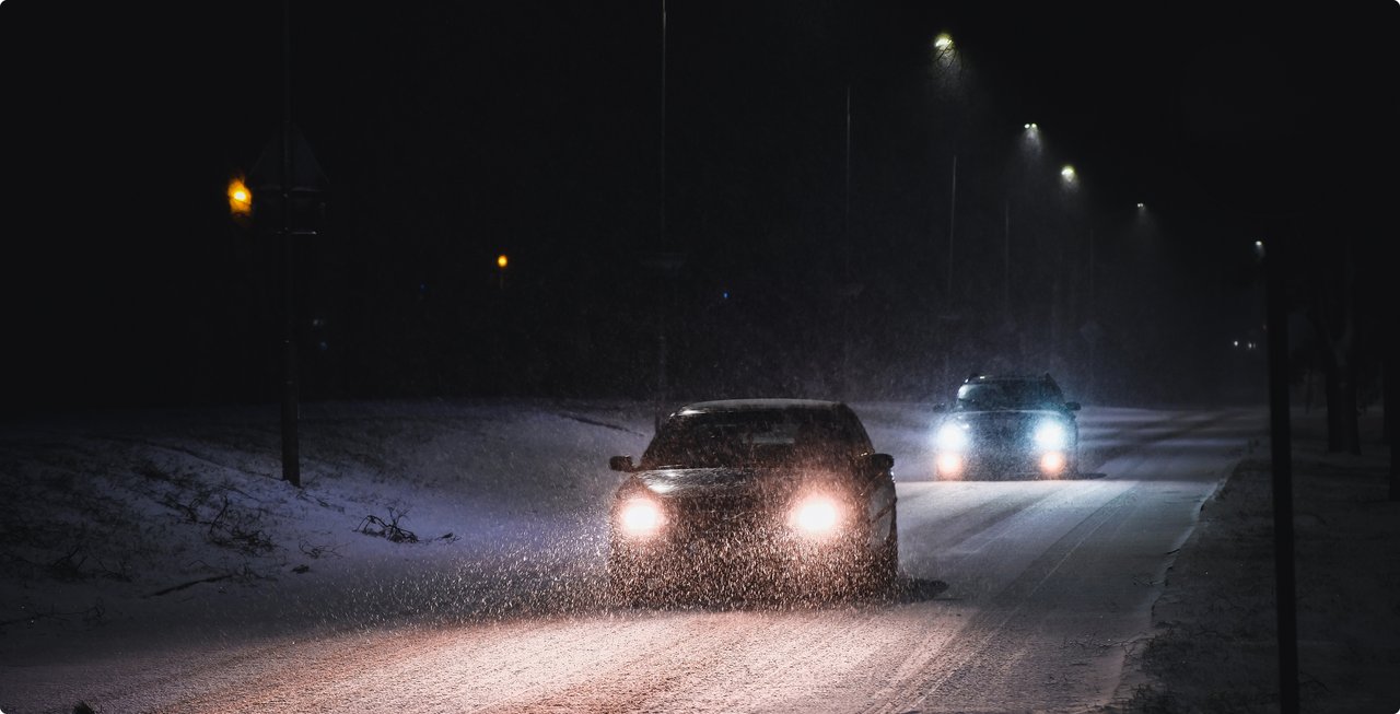 A car drives on a snow-covered road at night, its headlights illuminating the snow being kicked up in front of it. Other vehicles and streetlights are visible in the background.