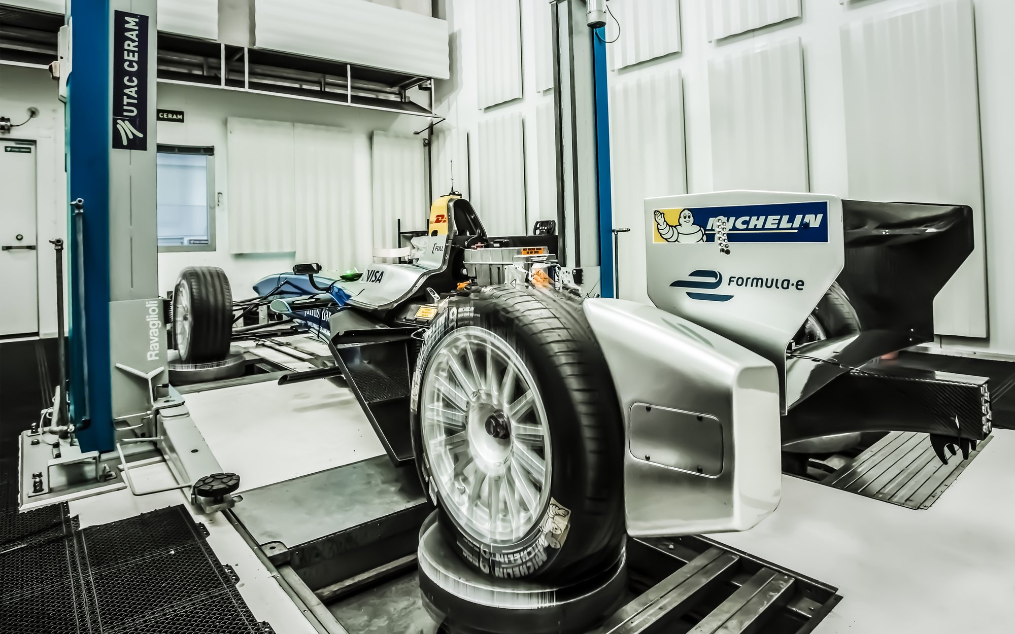 A vehicle chassis without a body is being tested in a laboratory setting with complex machinery and measurement devices. A wheel is prominently featured in the foreground.
