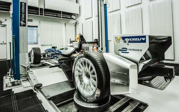 A vehicle chassis without a body is being tested in a laboratory setting with complex machinery and measurement devices. A wheel is prominently featured in the foreground.