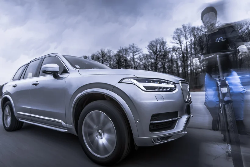 A silver SUV drives on a road, sharply focused in the foreground. To its right, a shadowy person or figure is visible, appearing partially blurred or transparent due to a blue light effect. The background shows trees and a cloudy sky, all in a slightly desaturated or monochrome style, interrupted by the blue effect on the figure.
