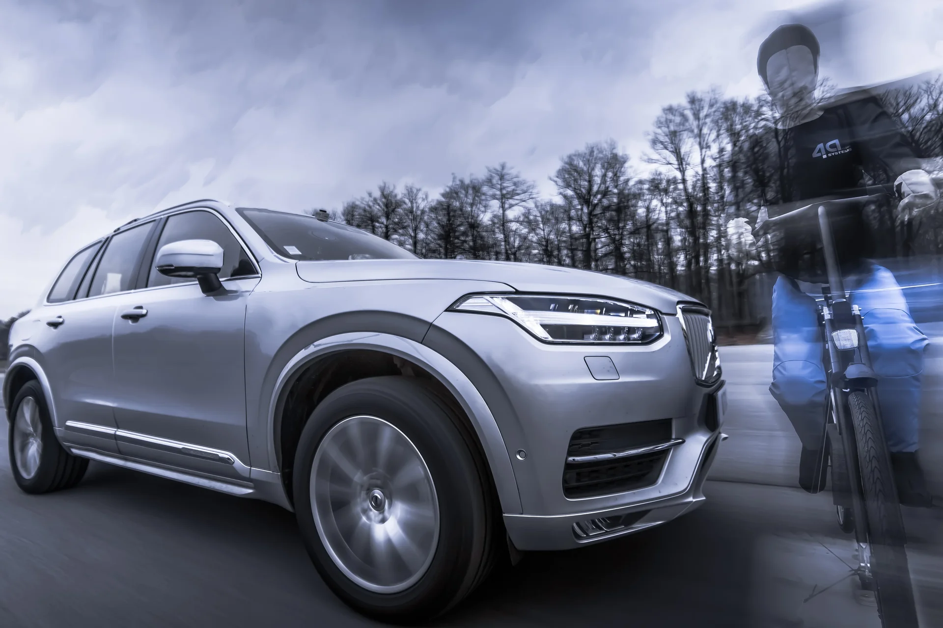 Un SUV argenté roule sur une route, net au premier plan. À sa droite, une personne ou une silhouette indistincte est visible, paraissant partiellement floue ou transparente en raison d'un effet de lumière bleue. L'arrière-plan montre des arbres et un ciel nuageux, le tout dans un style légèrement désaturé ou monochrome, interrompu par l'effet bleu sur la silhouette.